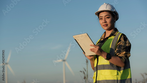 engineer in helmet with a clipboard on wind turbine on background. Asian woman in white helmet working with digital tablet at renewable energy farm. Female inspector controlling functioning of wind tu photo