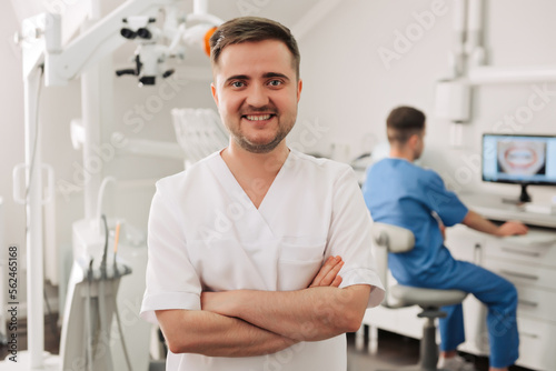 Portrait of a smiling dentist with arms crossed in dental clinic with his working assistant on the background