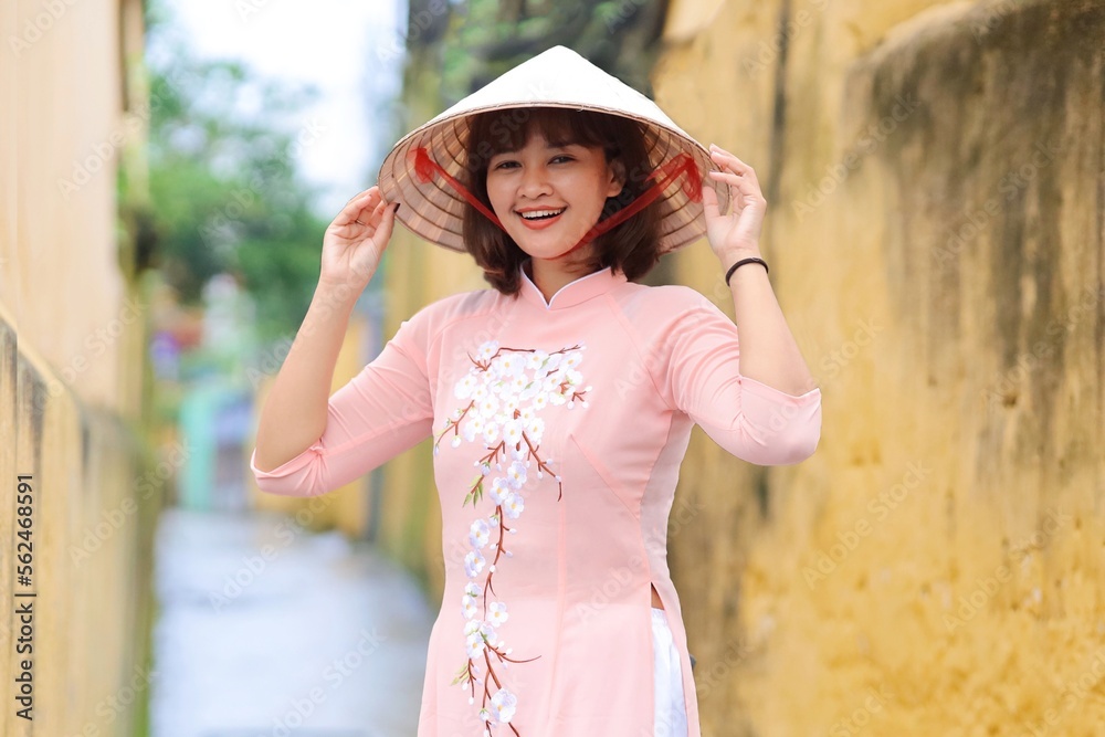 Smiling Asian woman in Traditional Vietnam dress standing over vintage yellow wall in accent town.