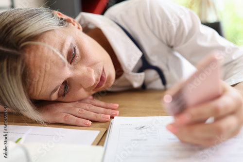 Tired female doctor laying on her work desk and looking at smartphone screen.