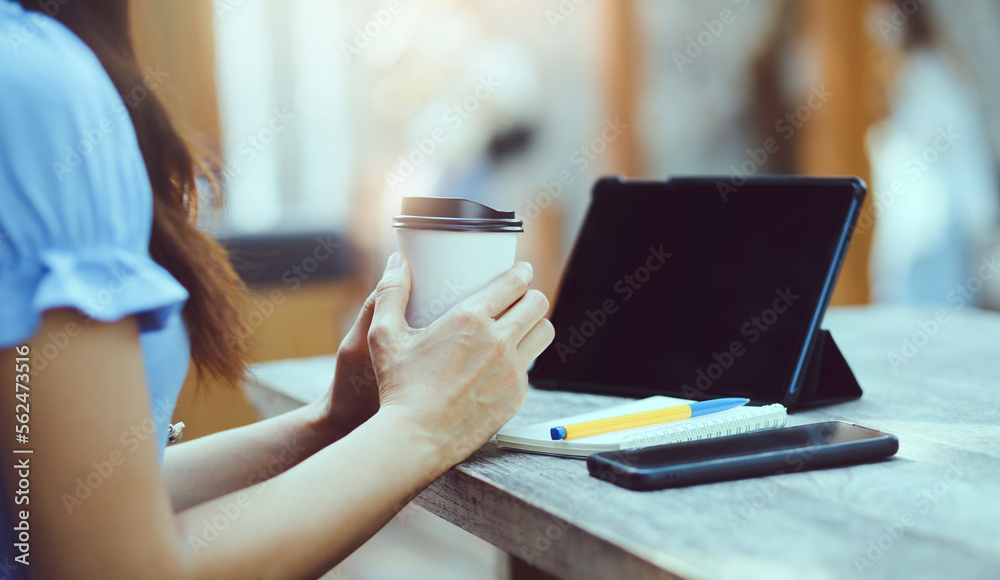 Asian freelance woman holding cup of hot coffee and working on tablet computer on wooden table at outdoor cafe. Entrepreneur woman working for her new business concept. Asian freelance woman holding cup of hot coffee and working on tablet computer on wooden table at outdoor cafe. Entrepreneur woman working for her new business concept.