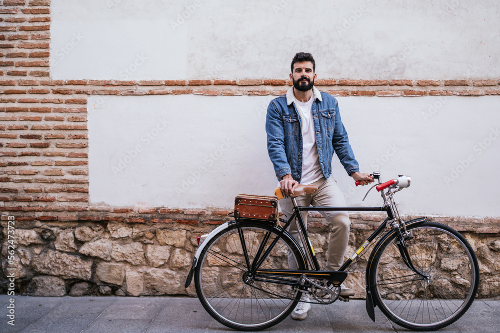 Obraz premium Young man in denim jacket holding his vintage classic bicycle while looking his mobile phone in front a brick wall.