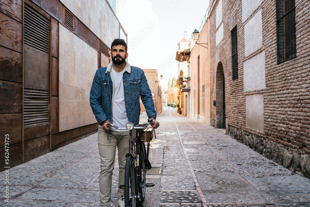 Young man in denim jacket pushing his vintage classic bicycle through the alleys of the city.