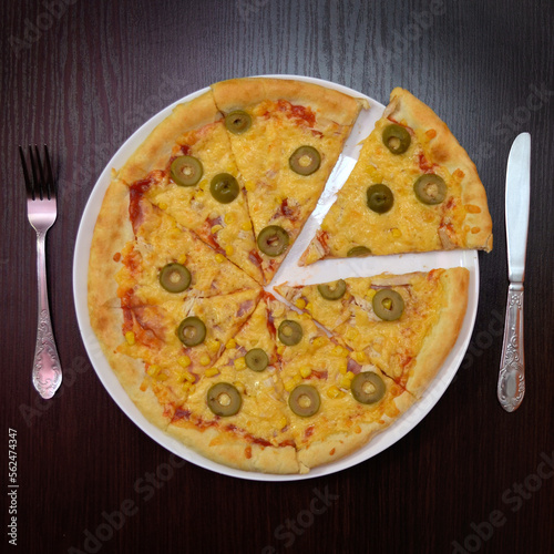 Delicious fresh cheese pizza with ham, olives on black table, surface, on white plate, fork, knife nearby, top view