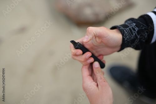 a guy of thirteen years old, holds a vape in his hands. A teenager smokes a disposable electronic cigarette while sitting by the sea