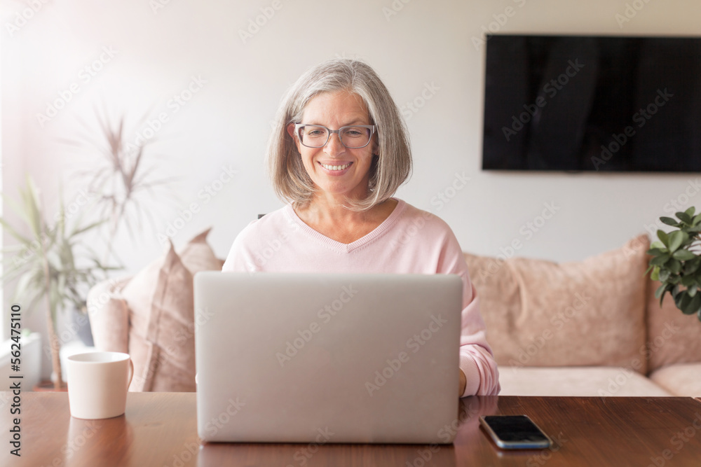 Happy middle age senior woman sitting at the table at home working using computer laptop Happy middle age senior woman sitting at the table at home working using computer laptop