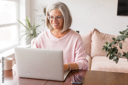 Happy middle age senior woman sitting at the table at home working using computer laptop photo