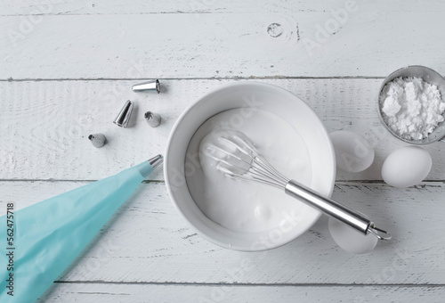 Royal icing or egg white glaze for decorating a cake or cookies. Served with whisk in a bowl and kitchen utensils such as piping bag  on white background table. Top view photo