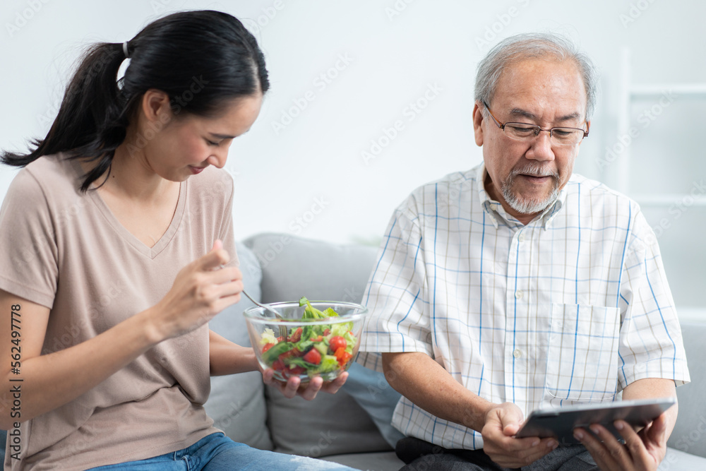 Young Asian daughter serving and feeding food to senior father while he using and playing on digital tablet in the living room. family love and support to elderly people. Young Asian daughter serving and feeding food to senior father while he using and playing on digital tablet in the living room. family love and support to elderly people.