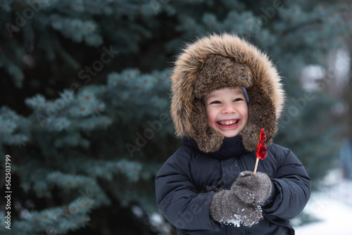 Photo Portrait of a beautiful little Russian boy in a hat with earflaps with a lollipop cockerel in winter in the park