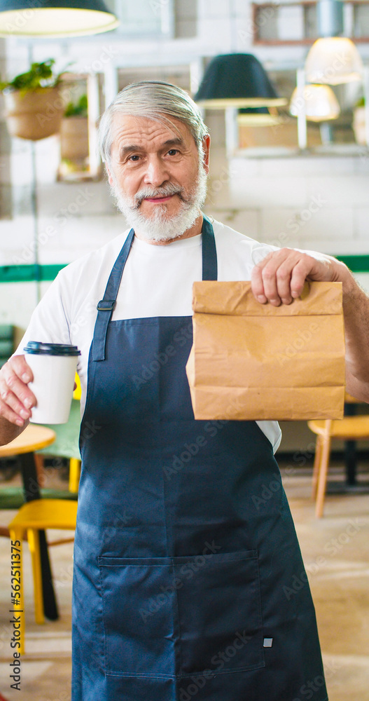Vertical footage of handsome happy senior positive Caucasian bearded man waiter in apron holding paper bag with take away order and coffee-to-go, looking at camera and smiling, business concept