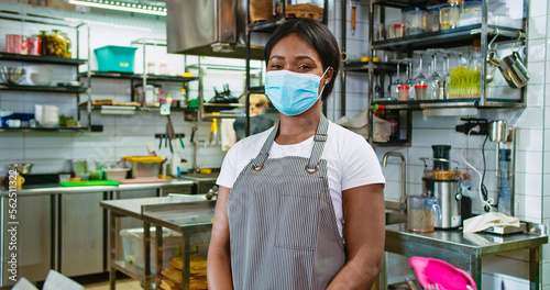 Portrait of young beautiful African American happy female in medical mask standing at workplace in restaurant at bar counter looking at camera and smiling. Work in kitchen, coronavirus concept