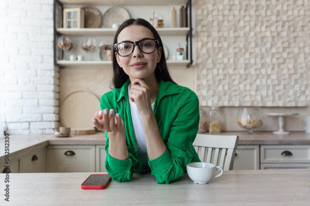 A young beautiful female blogger is sitting at home in the kitchen and talking online to the camera, explaining with her hands, smiling. A young beautiful female blogger is sitting at home in the kitchen and talking online to the camera, explaining with her hands, smiling.