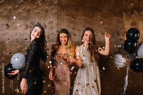 Charming adorable cute ladies in holiday dresses having fun and dancing under confetti. Three happy excited young women dancing with raised hands over dark background