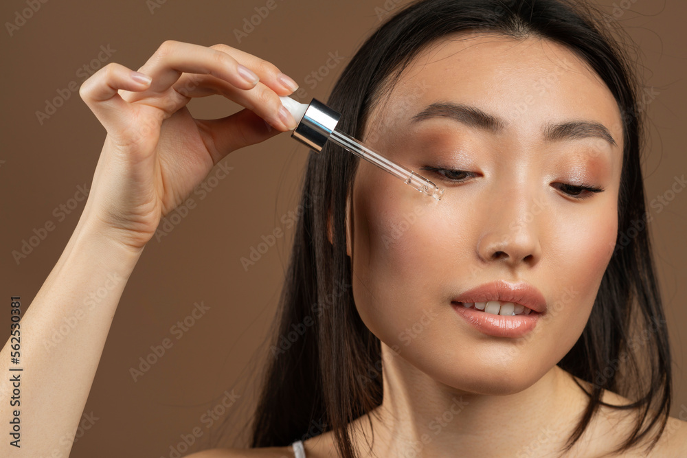 Pretty young asian lady using nourishing face serum after shower, enjoying organic beauty product, brown background