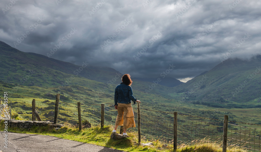Naklejka premium Young woman in a dress enjoys the unforgettable views of Killarney National Park, the dramatic skies and green mountains of the Ring of Kerry near the town of Killarney in County Kerry, 