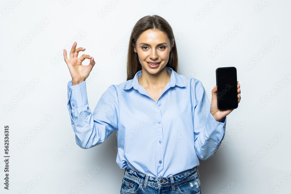 Smiling woman showing okay, mobile phone screen, recommending smartphone app, standing over white background Smiling woman showing okay, mobile phone screen, recommending smartphone app, standing over white background