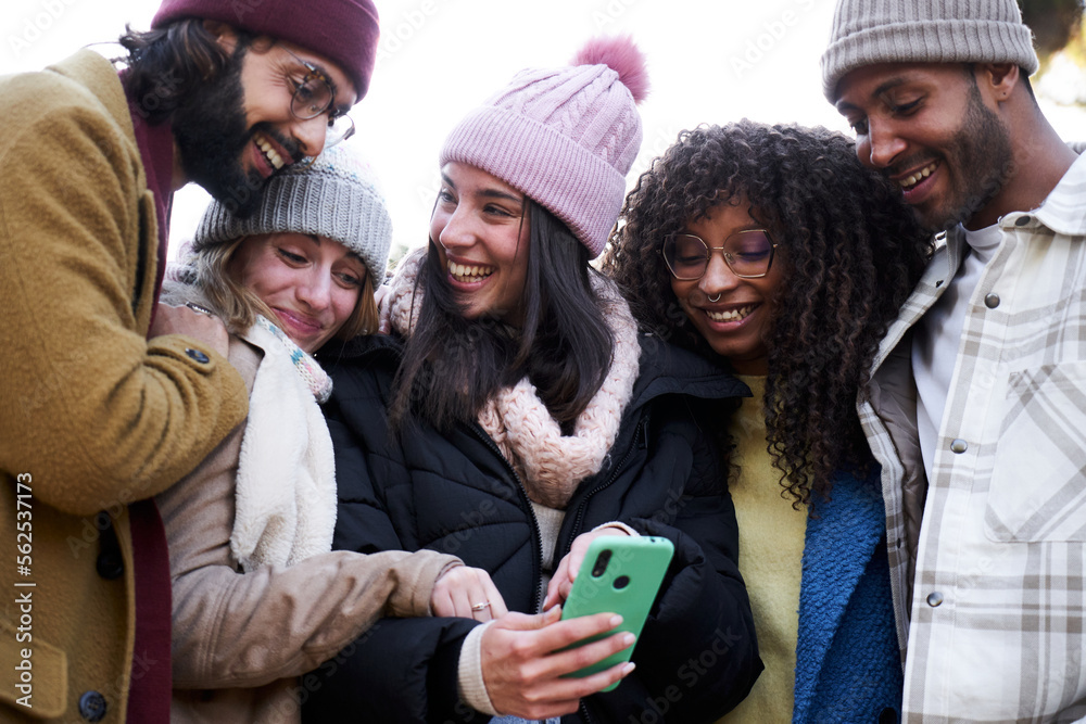 Group of multiethnic friends looking at mobile phone, laughing and sharing. Five people having fun outdoors. Concept of addicted to technology. Group of multiethnic friends looking at mobile phone, laughing and sharing. Five people having fun outdoors. Concept of addicted to technology.