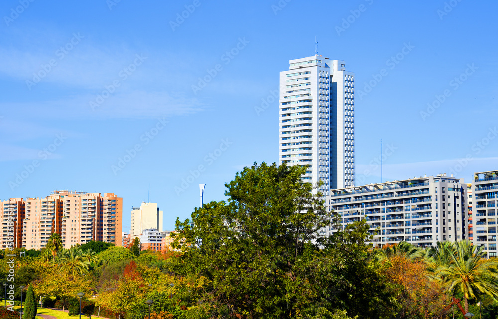 Green trees in park on background of facade buildings. Valencia Central Park with gardens on Turia River. Green trees in park. Urban landscape. Facade of a building and hotel. Fresh and clean air.