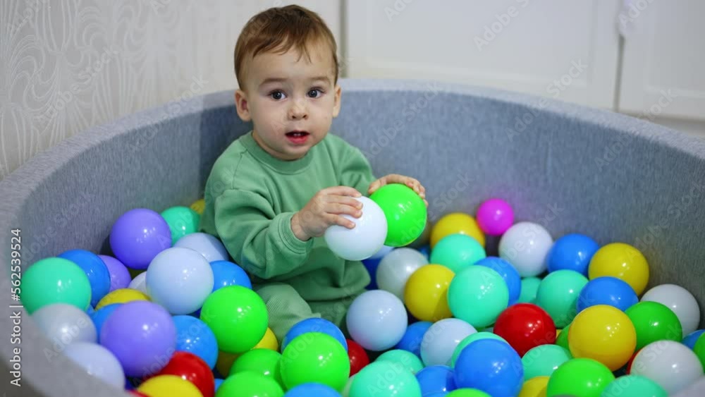 Adorable one year old child sits inside the dry pool. Baby boy holds the balls pulling them to his mouth. Top view. Adorable one year old child sits inside the dry pool. Baby boy holds the balls pulling them to his mouth. Top view.