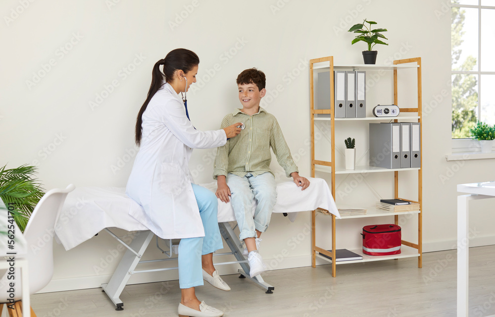 Child getting health checkup in exam room at doctor's office. Pediatrician sitting on medical couch together with school boy and examining his heart or lungs with stethoscope. Hospital visit concept Child getting health checkup in exam room at doctor's office. Pediatrician sitting on medical couch together with school boy and examining his heart or lungs with stethoscope. Hospital visit concept