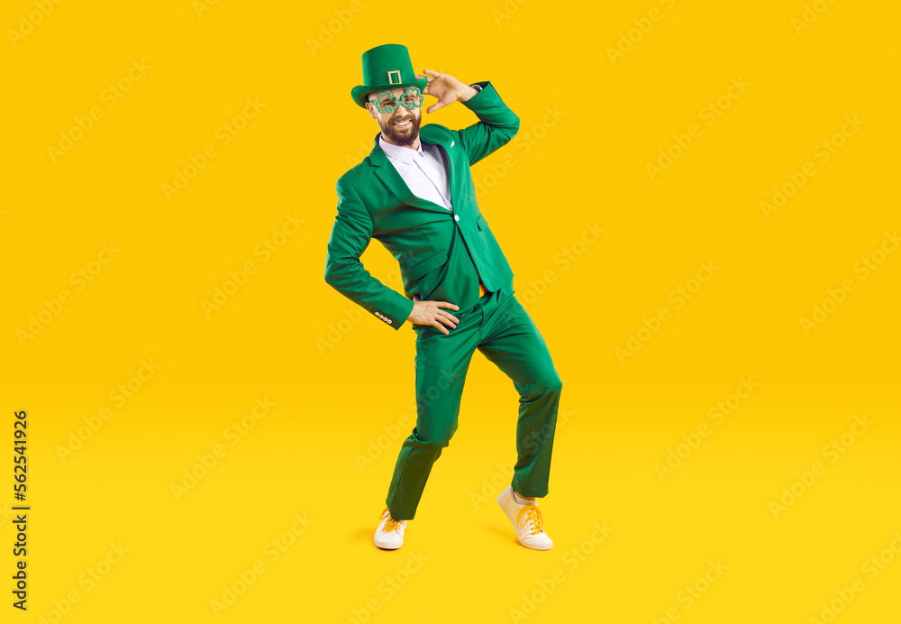 Happy St. Patrick's Day. Funny stylish man in green clothes celebrates St. Patrick's Day on orange background. Full length of young man in green suit with hat and party glasses dancing and having fun.