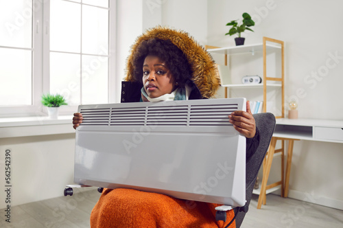 Photographie Portrait of a young disappointed worried frozen african american woman sitting in a warm winter coat with a hood on the grey chair at home and holding electric heater
