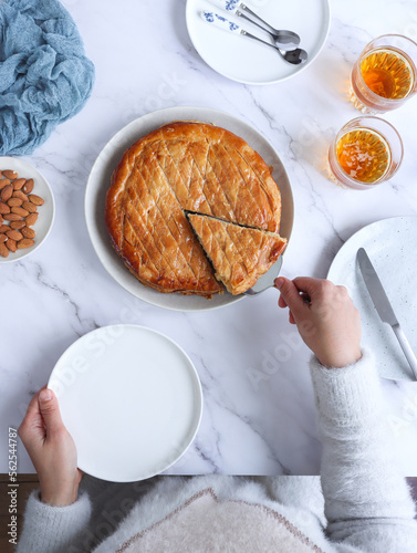 Top view of French galette des rois with cider, hands holding plate and grabbing a piece with handle cake slicer. It's made with puff pastry and creamy almond filling roll in circles shape.  photo