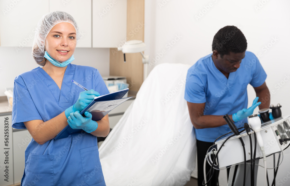 Obraz premium Young female cosmetologist in blue overall filling out medical form while male assistant preparing equipment for procedures in aesthetic medicine office