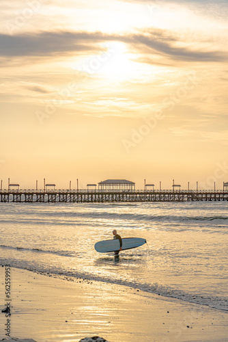 surfer on the beach under a beautiful sunset in front of the pier with big sea waves man on the beach on an incredible sunset with the surfboard, walking along the shore of the beach