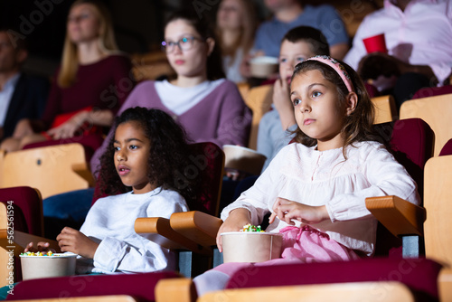 Preteen girl with her mulatta mate sitting in comfortable chairs in cinema, eating popcorn and absorbedly watching movie