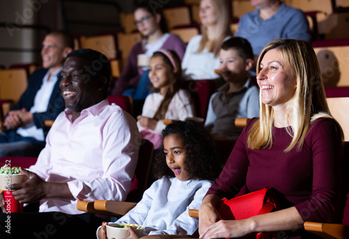Portrait of laughing european woman watching funny movie in cinema with her african husband and preteen daughter