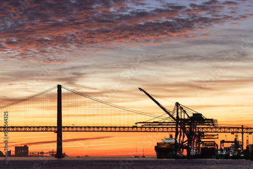 Industrial cranes, a suspension bridge, and a tourist liner sailing along the river at sunset
