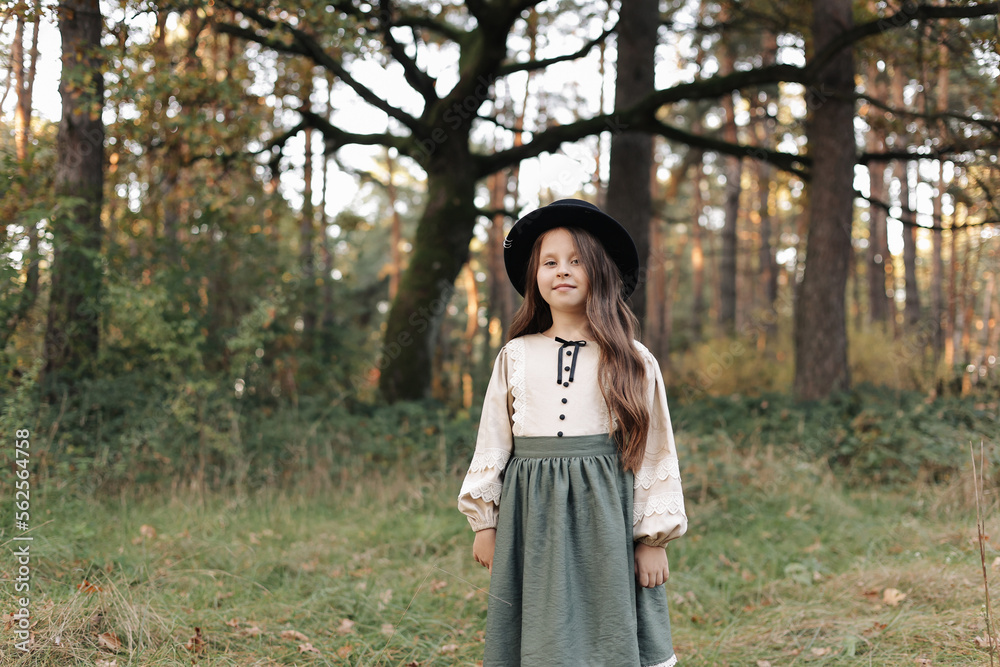 Portrait of little caucasian girl looking at camera standing outdoors. Stylish adorable smiling child in black hat enjoying childhood testimonial concept at green park background.