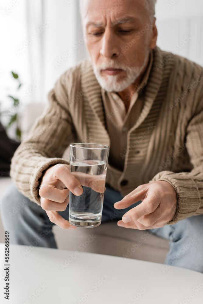 blurred man with parkinson syndrome and tremor in hands sitting and holding glass of water at home.