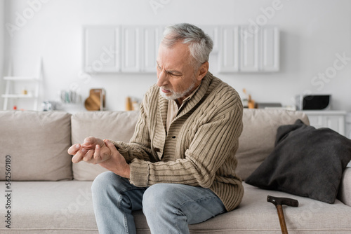 aged man with parkinson syndrome suffering from tremor in hands while sitting on sofa near walking cane. photo