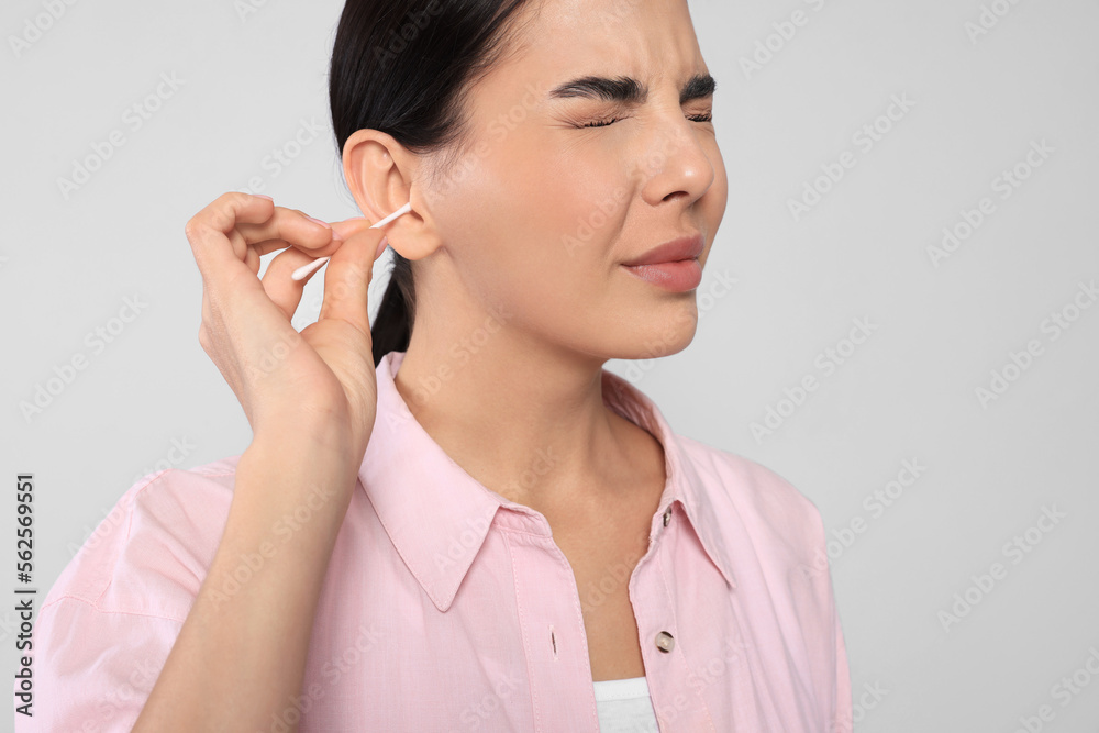 Young woman cleaning ear with cotton swab on light grey background, closeup