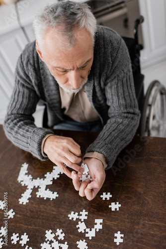 high angle view of man with parkinsonian syndrome sitting in wheelchair and holding elements of jigsaw puzzle in trembling hands.