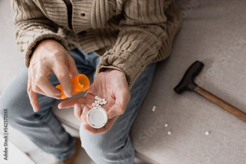 cropped view of aged man suffering from parkinsonism and holding container with pills while sitting on couch.