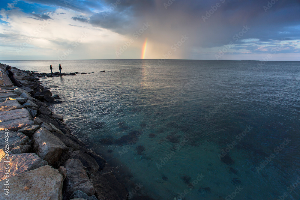 Rainbow and cloudy sky over silhouettes of two people fishing on coastal rocks, Massachusetts, USA Rainbow and cloudy sky over silhouettes of two people fishing on coastal rocks, Massachusetts, USA