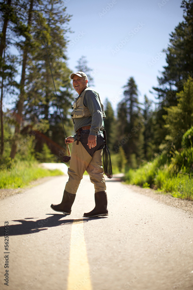A fisherman holding a fly rod walks away from the camera, looking back and smiling. A fisherman holding a fly rod walks away from the camera, looking back and smiling.