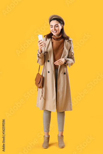 Young woman in coat using mobile phone on yellow background