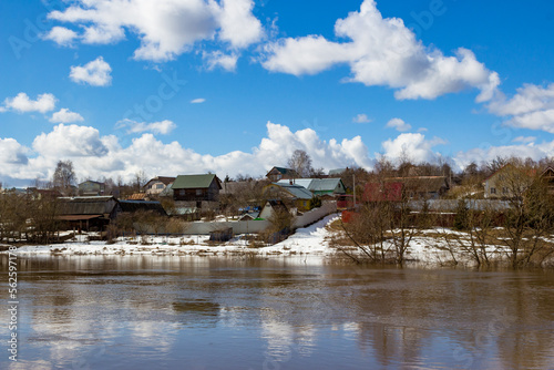Rural buildings on a hill beyond the river against a blue sky with clouds, a river overflowing in flood