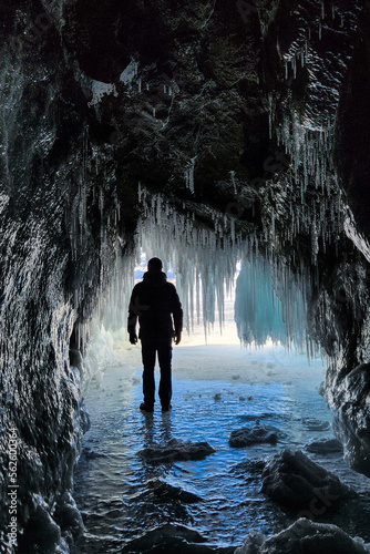 A man is hiking in a beautiful ice cave. A man traveling in winter, standing on a frozen lake with an ice cave. A man explores an amazing glacier cave . Selective focus.  © Nikita