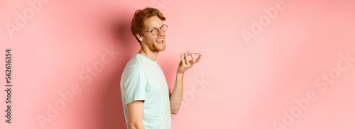 Handsome young man standing in profile and talking on speakerpone, record voice message, turn head at camera and smiling pleased, standing over pink background