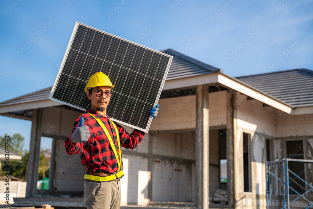 Asian technician holding solar panel with thumbs up showing confidence in front of unfinished house at construction site. Concept of alternative and renewable energy. Asian technician holding solar panel with thumbs up showing confidence in front of unfinished house at construction site. Concept of alternative and renewable energy.