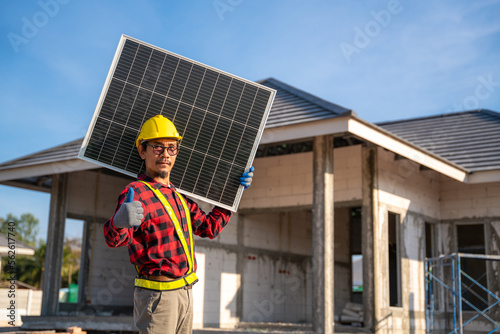 Asian technician holding solar panel with thumbs up showing confidence in front of unfinished house at construction site. Concept of alternative and renewable energy. © tong2530