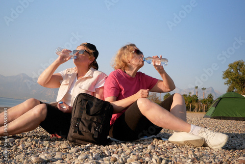 Two women tourists drinking water from bottle on sea beach of tent background in sunny day. Tourist tired.