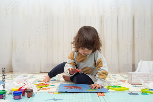 Little girl painting with finger paint a handprint on a blue cardboard in a playroom. photo