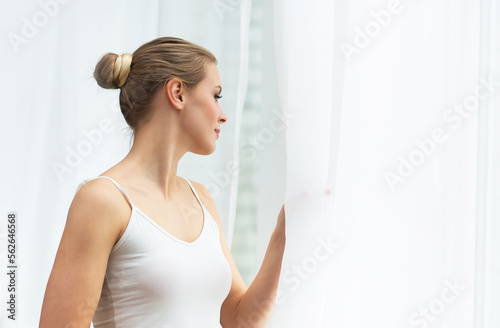 Young woman in white underwear is standing in front of the window and looking into it. Girl at home in the living room.